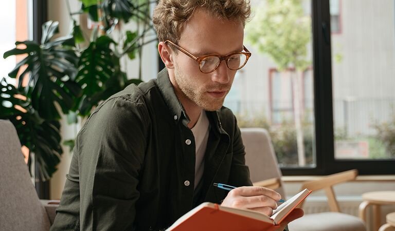 Man with red glasses and a green button up shirt writes in a red notebook. His office is bright and open, with a large fern.